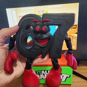 May include: An articulated figure in the shape of the number 67. The figure is black with red accents, including boxing gloves for hands and feet, and a smiling face. It stands on a green box. The figure is approximately 15 cm tall.