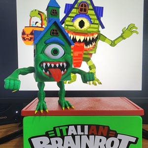 Pode incluir: Uma caixa verde e vermelha com o texto "ITALIAN BRAINROT" em branco. A caixa tem uma parte superior vermelha e laterais verdes. Uma casa monstro verde com um olho grande e uma língua está em cima da caixa. Outra casa monstro está no fundo.