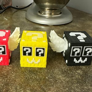 May include: Three colourful, winged cube-shaped objects. The cubes are red, yellow, and black, each with a white question mark on top and two question marks below. The cubes have white wings and a pixelated mouth.