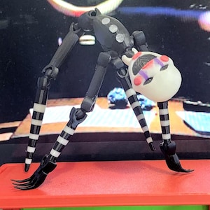 May include: A black and white articulated figure of a character with a white head and a painted face. The figure has long, thin legs with black and white stripes. The figure is posed on a red surface.