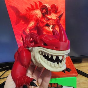 Pode incluir: Uma figura vermelha e branca impressa em 3D de uma criatura com chifres e dentes afiados. A figura é segurada em frente a uma ilustração vermelha da mesma criatura. A base é verde com o texto "AN TAROT".