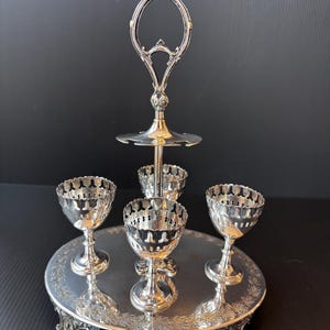 Könnte beinhalten: Ein silberfarbener, mehrstöckiger Servierständer mit vier Eierbechern. Der Ständer hat einen runden Sockel mit floralen Details, eine zentrale Stütze und einen dekorativen Griff. Die Eierbecher haben ein filigranes Design.