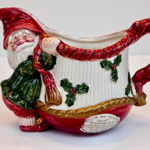 May include: A ceramic Christmas creamer featuring a Santa Claus figure. The creamer has a red handle and base, a white body with vertical lines, and is decorated with holly leaves. The Santa figure wears a red hat and green coat.