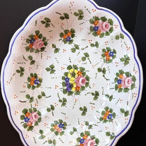Puede incluir: Plato de cerámica blanco con borde festoneado y borde azul. El plato está decorado con coloridos motivos florales, que incluyen flores rosas, naranjas, amarillas y azules, y hojas verdes.