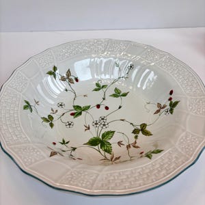 Puede incluir: Bol de cerámica blanco con borde decorativo y ribete verde. El cuenco presenta un diseño floral con hojas verdes, bayas rojas y flores blancas. El borde tiene un patrón de tejido de cesta.
