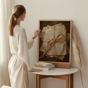 Romantic vintage love letters wall art shown in a styled home interior with a woman gently placing a framed print on a round table. The artwork features antique handwritten letters tied with a heart ribbon, aged paper textures, and subtle red heart details. Soft neutral tones, natural daylight, minimal decor, stacked books, and dried pampas grass create a calm, elegant atmosphere. Ideal for Valentine wall art, romantic printable decor, and vintage-inspired living spaces.