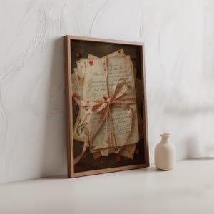 May include: A framed print featuring a stack of aged letters tied with a ribbon adorned with red hearts. The letters have handwritten script. Small red heart-shaped embellishments are scattered around the frame. A small, off-white vase sits to the right.