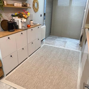 May include: A rectangular beige rug with a white border is placed on a tiled floor. The rug has a woven texture and is in a kitchen setting. Cabinets, shelves, and appliances are visible in the background.