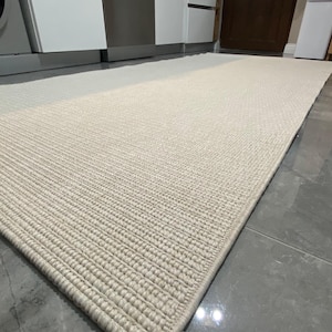 Puede incluir: Una alfombra rectangular tejida de color crema con un patrón acanalado texturizado. La alfombra está colocada sobre un suelo de baldosas grises y se ve el borde de la alfombra. La alfombra está en un entorno de cocina.