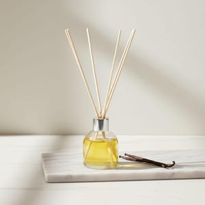 Pode incluir: Um difusor de vidro transparente com tampa prateada, preenchido com líquido amarelo e varetas de madeira. Uma vagem de baunilha repousa sobre uma superfície de mármore branco, sugerindo um aroma de baunilha. Para perfumar a casa.