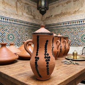 Peut inclure: Une collection de poteries en terre cuite sur une table en bois. La pièce maîtresse est une grande cruche avec un couvercle et une anse, ornée de motifs de points noirs. Autres pièces : tagines et cruches.