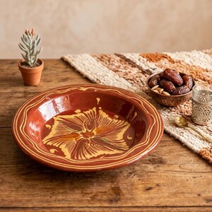 Peut inclure: Un bol en céramique de couleur terre cuite avec un motif floral jaune et orange. Le bol a un bord festonné et une ligne jaune décorative autour du bord. Le bol est sur une table en bois, avec une petite plante en pot et un bol de dattes en arrière-plan.