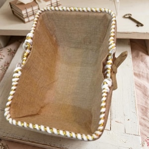 Peut inclure: Panier rectangulaire en toile de jute avec une bordure en corde blanche et dorée. Le panier a des poignées en bois et est posé sur un plateau en bois blanc. Un petit tas de livres avec un ruban crème est en arrière-plan.