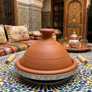 May include: A terracotta tagine pot with a conical lid and a metal base, set on a mosaic tile table. The pot has wooden handles and is surrounded by a traditional Moroccan setting with colorful cushions and ornate architecture.