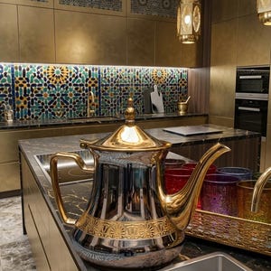 Peut inclure: Une théière argentée et dorée aux détails complexes est posée sur un comptoir de cuisine. La cuisine comprend des armoires dorées, une crédence carrelée colorée et des lanternes décoratives suspendues. Un plateau de verres colorés est également visible.