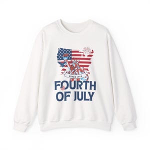 Peut inclure: Un sweat-shirt blanc à manches longues avec une impression graphique de la Statue de la Liberté, un pygargue à tête blanche, des feux d'artifice et le drapeau des États-Unis en forme de pays. Le texte indique SINCE 1776 et FOURTH OF JULY.