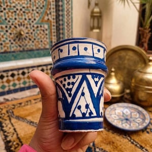 May include: A blue and white ceramic cup with a geometric design. The cup has a cylindrical shape with a flared rim and is decorated with blue patterns on a white background. The cup is held in a hand.