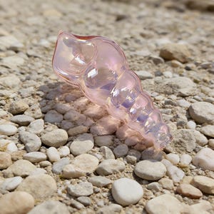 May include: A translucent, iridescent pink glass seashell-shaped object. The glass has a pearlescent sheen, reflecting light with hints of purple and gold. The object is resting on a bed of small, light-colored stones.
