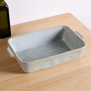 Peut inclure: Un plat à four rectangulaire en céramique bleu clair, avec des coins arrondis et de petites poignées sur les côtés. Le plat a une texture tachetée et repose sur une surface en bois clair. Une bouteille d'huile d'olive est partiellement visible.