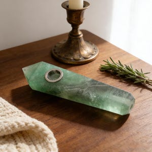 May include: A green fluorite pipe with a silver metal bowl. The pipe is rectangular with angled edges. The pipe is on a wooden surface. A brass candlestick and a sprig of rosemary are in the background.