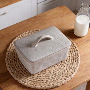 Könnte beinhalten: Ein gesprenkelter, grauer Keramik-Butterbehälter mit einem gebogenen Griff auf dem Deckel, der auf einem runden, gewebten Platzset liegt. Eine Glasflasche mit Milch steht daneben auf einem Holztisch. Der Butterbehälter ist rechteckig.