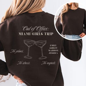 May include: A dark brown sweatshirt with the text "Out of Office: MIAMI GIRLS TRIP" and the names EMILY, ASHLEY, MADISON, and JESSICA. The design includes two cocktail glasses and the phrases "No plans. No stress. No regrets."