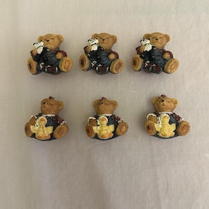 May include: A set of six small bear figurines arranged in two rows. The top row features three brown bears wearing dark blue overalls and holding small panda toys. The bottom row features three brown bears in dark blue overalls holding yellow ducks.