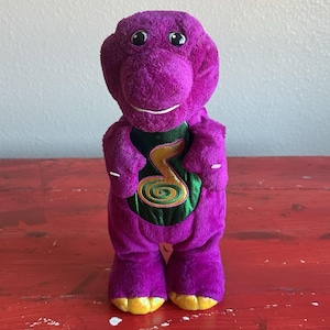 May include: A plush purple dinosaur toy with a green vest featuring a gold musical note design. The toy has yellow feet and a smiling face. The background is a red surface.