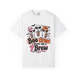 May include: A white short-sleeve t-shirt featuring a graphic of three cartoon ghosts wearing sunglasses and holding iced coffee cups. The text reads Boo Crew Runs on 7 Brew, surrounded by small bats, pink hearts, and sparkles in orange and pink.