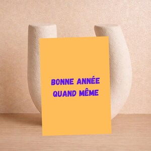 May include: An orange card with the text "BONNE ANNÉE QUAND MÊME" in blue, set against a neutral background. The card is propped up by a white, abstract ceramic sculpture. The overall aesthetic is minimalist and modern.