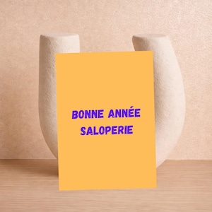 May include: An orange card with the text "BONNE ANNÉE SALOPERIE" in blue lettering. The card is propped up against a white, abstract ceramic sculpture. The background is a light peach color, and the surface is a light brown.