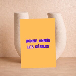 May include: A greeting card with the text "BONNE ANNÉE LES DÉBILES" in blue on an orange background. The card is propped up against a white, abstract sculpture. The background is a light peach color.