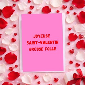 May include: A pink greeting card with red text that reads "JOYEUX SAINT-VALENTIN GROSSE FOLLE" is surrounded by red and white rose petals and heart-shaped decorations. A wrapped gift with a red ribbon is in the bottom right corner.