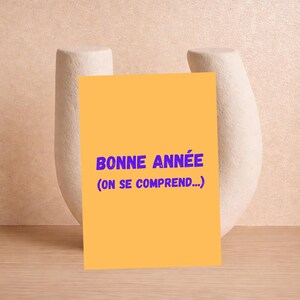 May include: A yellow card with the text "BONNE ANNÉE (ON SE COMPREND...)" in purple, placed in front of a white, abstract ceramic vase. The background is a light peach color, and the card is resting on a wooden surface.