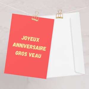 May include: A red card with gold text reading "JOYEUX ANNIVERSAIRE GROS VEAU" is clipped to a string with a gold binder clip, next to a white envelope. The card is the focal point.