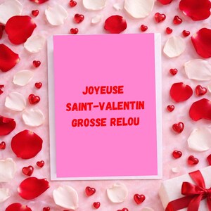 May include: A pink greeting card with the text "JOYEUX SAINT-VALENTIN GROSSE RELOU" in red, surrounded by red and white rose petals, heart-shaped candies, and a wrapped gift with a red ribbon. The card is centered.