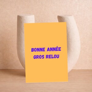 May include: A yellow card with the text "BONNE ANNÉE GROS RELOU" in blue. The card is propped up in front of a white, abstract sculpture against a neutral background. The text is in French.