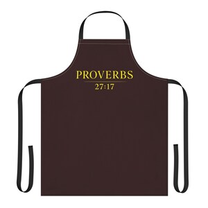 May include: A dark brown apron featuring black neck and waist ties. The text PROVERBS 27:17 is printed in yellow across the upper chest area. The apron has a standard rectangular body shape with a curved neckline and long black fabric ties.
