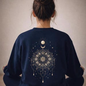 Könnte beinhalten: Marineblaues Sweatshirt mit goldenem Mandala-Design auf dem Rücken, mit Sonne, Mond und Sternen. Das komplizierte Muster umfasst ein Blumenzentrum und hängende Perlenakzente. Die Person trägt eine dunkelblaue Hose.