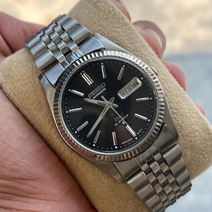 May include: A stainless steel Seiko automatic wristwatch with a black dial. The watch features a day and date display, a fluted bezel, and a silver-tone bracelet. The watch is displayed on a beige surface.