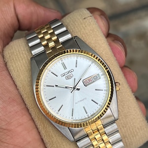 May include: A Seiko 5 automatic wristwatch featuring a silver dial with baton indices, a gold-tone fluted bezel, and a two-tone stainless steel link bracelet. The dial displays the Seiko 5 logo, the word AUTOMATIC, and a day-date window at 3 o'clock.