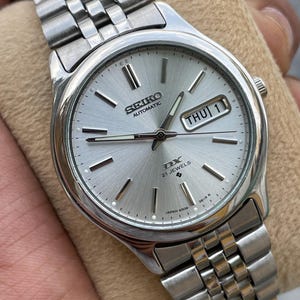 May include: A silver Seiko DX automatic wristwatch with a silver dial and a stainless steel band. The watch face displays the day and date. The watch is on a beige surface.