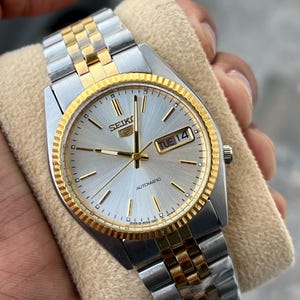 May include: A silver and gold-toned Seiko 5 automatic watch with a fluted bezel and a silver dial. The watch features a day and date display and a two-tone stainless steel and gold-toned bracelet.