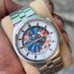 May include: A stainless steel wristwatch with a silver-tone metal link bracelet. The watch face features a colorful map of the world with blue oceans and orange landmasses, a 24-hour scale, and the text EDOX GEOSCOPE printed on the white dial.