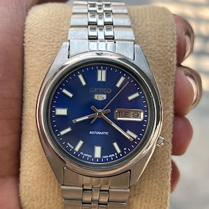 May include: A stainless steel Seiko 5 automatic watch with a blue dial. The watch features a day and date display, luminous hands and markers, and a silver-tone bracelet. The watch is marked "Made in Japan".