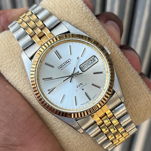 Pode incluir: Um relógio de pulso automático Seiko DX prateado e dourado. O relógio apresenta um mostrador prateado com detalhes dourados, um visor de dia e data e uma pulseira bicolor em aço inoxidável e dourado. O relógio é segurado contra um fundo bege.