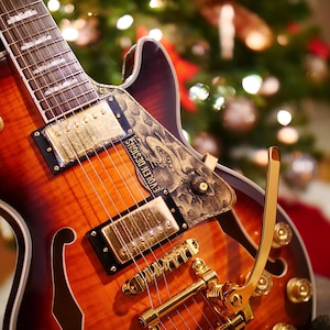 Puede incluir: Una guitarra eléctrica con acabado sunburst, herrajes dorados y un golpeador decorativo. La guitarra presenta un cuerpo tallado con agujeros en forma de f y el texto "EVOLEM DESIGNS" en el golpeador. El fondo está borroso, con un árbol de Navidad visible.