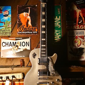 Puede incluir: Una guitarra eléctrica plateada con clavijero negro y marcadores de traste blancos se exhibe contra una pared de madera. Carteles metálicos vintage con texto e ilustraciones rodean la guitarra, incluyendo "Pepsi-Cola", "Indian" y "Champion".