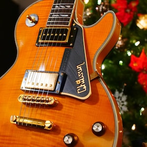 Könnte beinhalten: Nahaufnahme einer Vintage-E-Gitarre mit geflammter Ahorndecke. Die Gitarre verfügt über goldfarbene Hardware, einschließlich Steg, Tonabnehmer und Stimmwirbel. Das "Gibson"-Logo ist sichtbar. Der Hintergrund ist ein verschwommener Weihnachtsbaum.