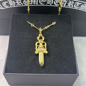 May include: A gold pendant necklace with a dagger design, set with clear gemstones. The necklace is presented in a black box, with the brand name "CHROME HEARTS" visible on the box.
